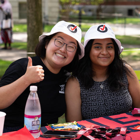 Two students smile for the camera wearing Harvard Votes Challenge hats, while sat in Harvard Yard at a table.